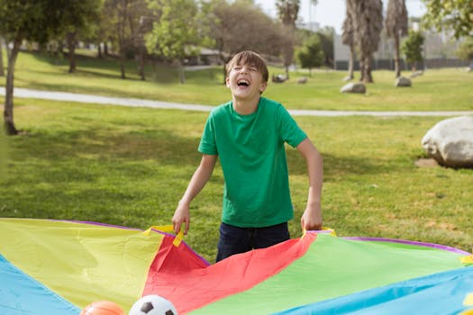 Laughing boy in green shirt playing with colorful parachute outdoors on a sunny day.