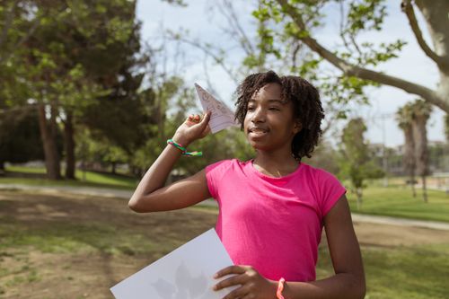 Free Teen girl with afro hair enjoys playing with a paper airplane in a sunny park. Stock Photo