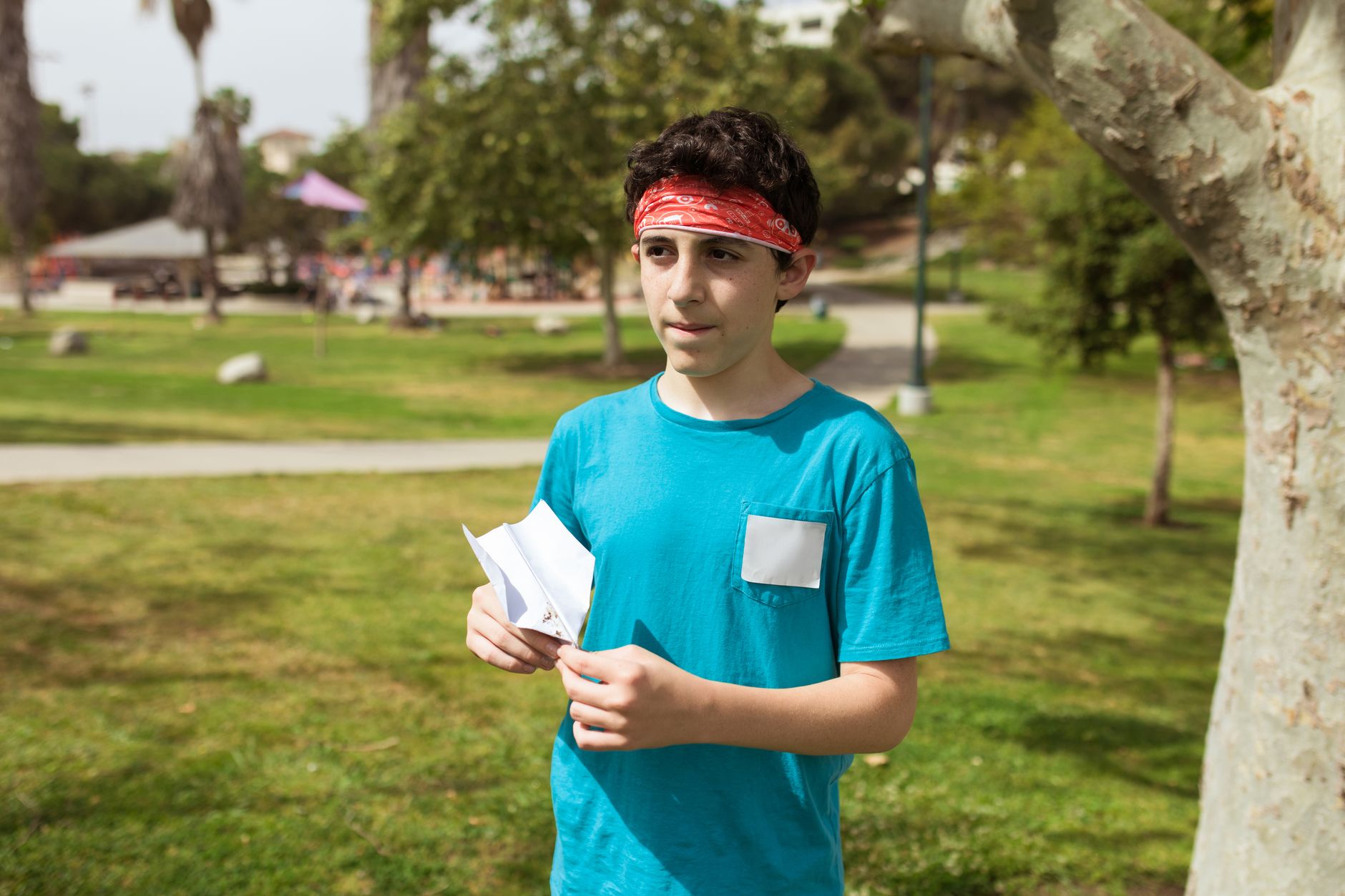 A young boy with a bandana holding a paper airplane in a sunny park setting.