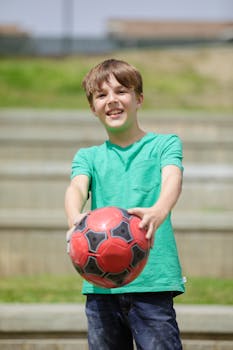 Young boy smiling, holding a soccer ball outdoors with blurred steps background