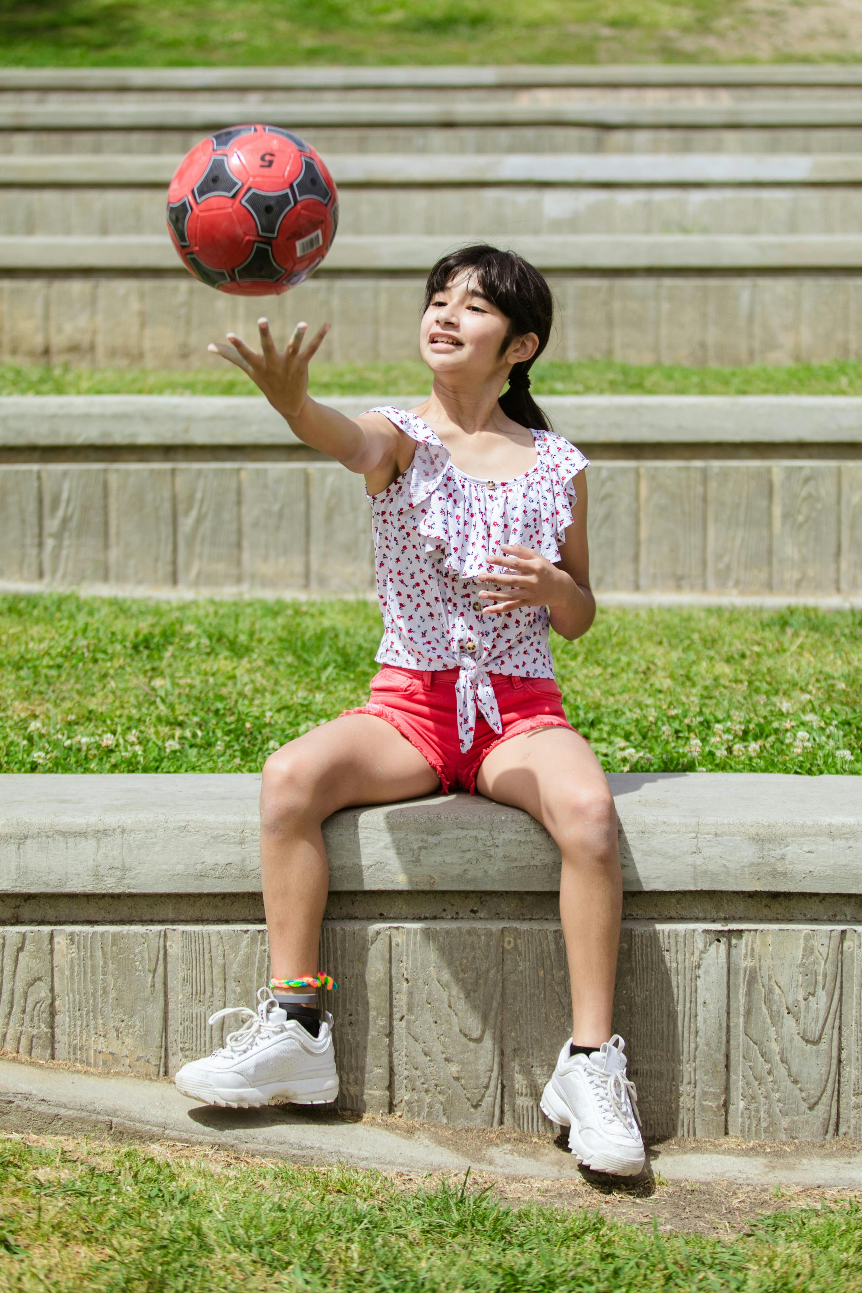 a girl in floral top sitting on a concrete bench while playing a ball