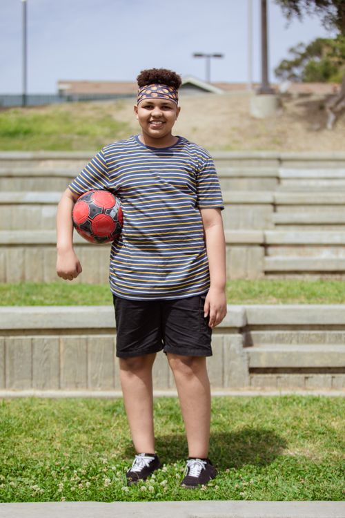 Free Happy boy in a striped shirt holding a soccer ball, standing outdoors on a sunny day. Stock Photo