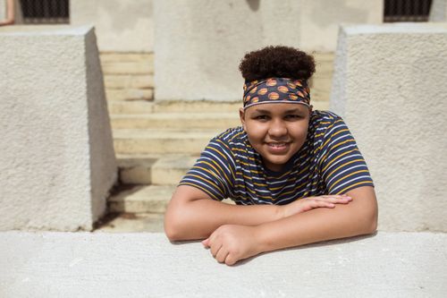 Free A young boy with curly hair smiling and leaning on a concrete structure outdoors. Stock Photo