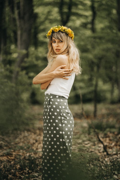 Side view of charming young female wearing casual summer clothes and yellow wildflower wreath on head embracing herself in green forest