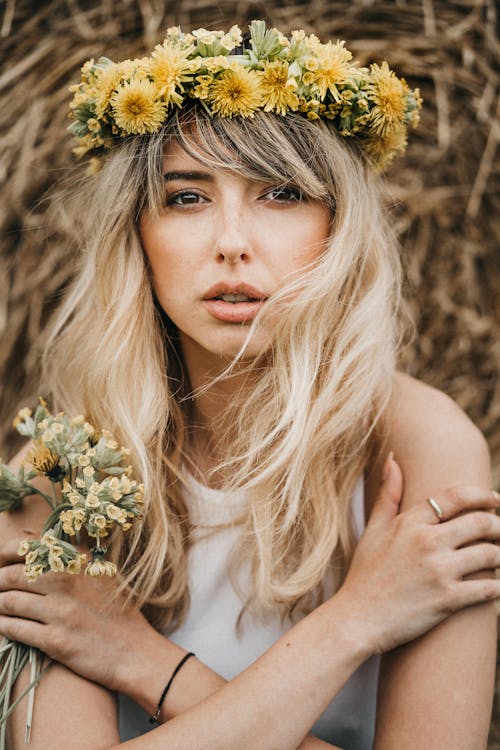 Tranquil female with blond long hair and wearing wreath of dandelions hugging for hands with bouquet of field flowers