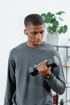 Young man in gray sweater lifting a dumbbell indoors, focusing on fitness routine.