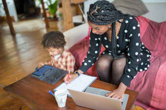 A mother writes notes on paperwork beside a laptop at a wooden coffee table while her young child uses a tablet next to her
