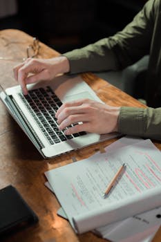 Focused person typing on laptop with notes and papers beside on wooden table.