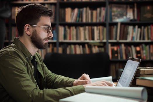 Focused young man in library working on laptop, typing amid shelves of books.