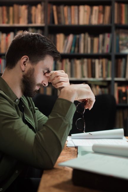 A tired man with glasses in a library, holding his head, symbolizing stress or fatigue. Overcome Academic Procrastination And Chronic: Overcome Academic Procrastination & Chronic Distraction: 5+ Tips - illustration 2