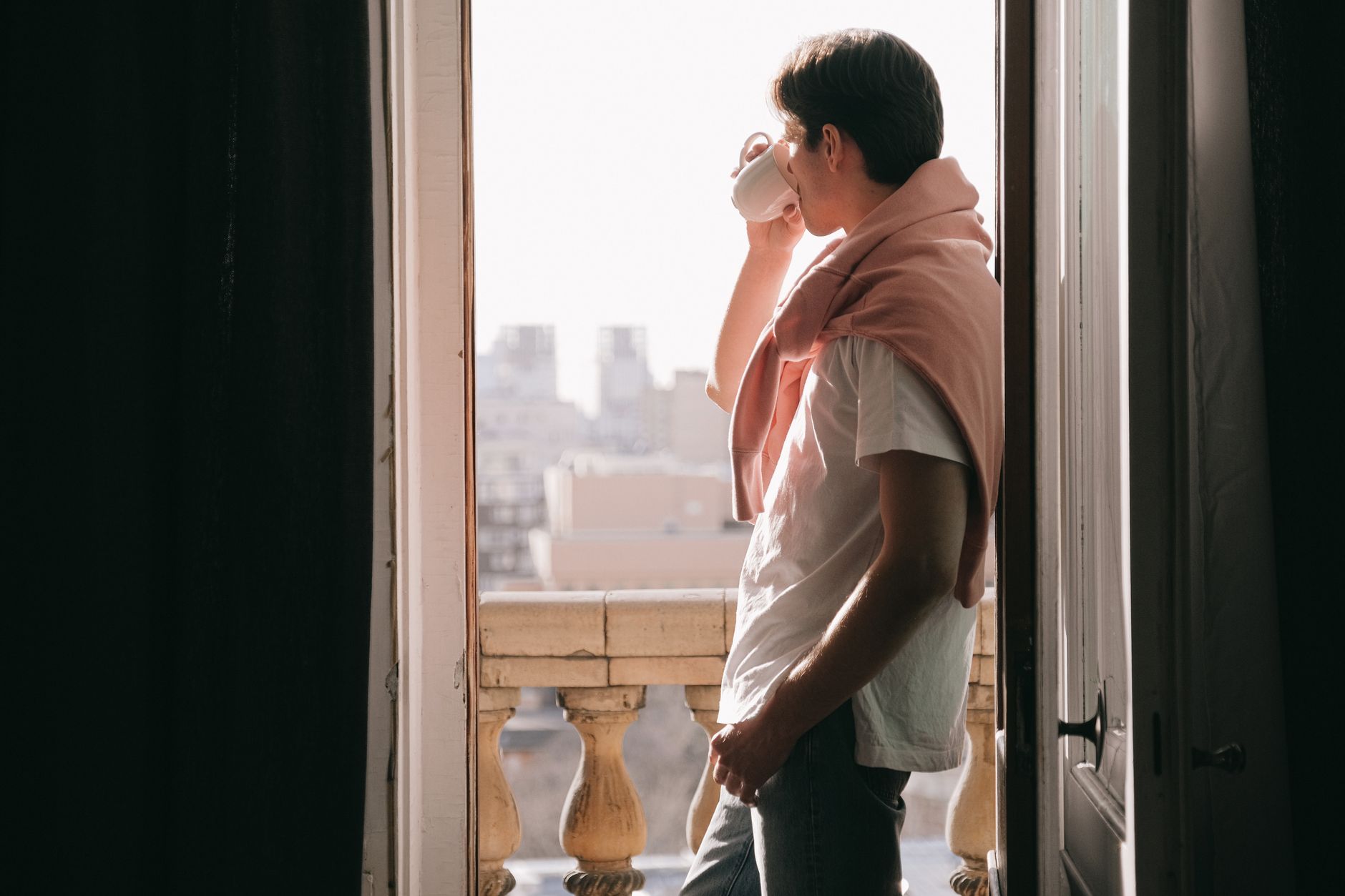 A man sipping coffee while standing in a doorway overlooking an urban balcony.