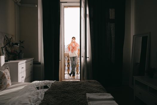 A man stands on a balcony enjoying coffee, morning light streaming into a cozy bedroom.