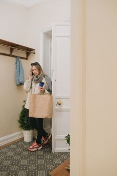 Caucasian woman holding coffee and paper bag near indoor doorway, making a phone call.