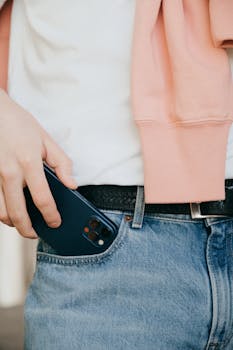 Close-up of a person placing a phone into denim jeans pocket with a pink jacket draped.