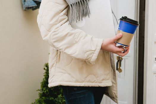 A person in a casual jacket holding a laptop, keys, and coffee cup outdoors.