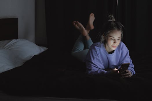 A woman lies in bed at night, using her smartphone with a relaxed expression.