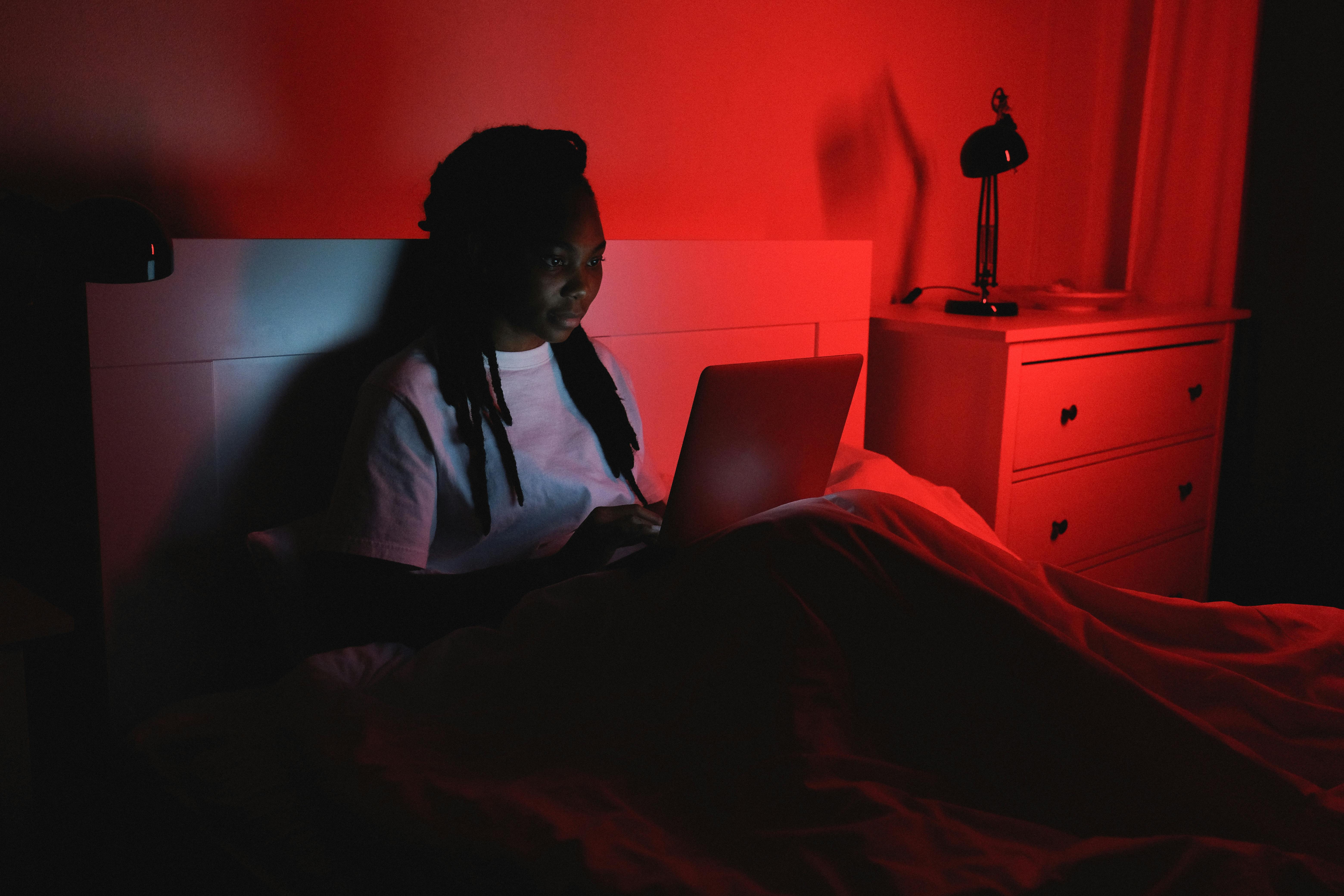 A woman sits in bed using a laptop, illuminated by red ambient light.