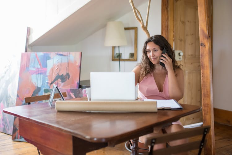 Woman Sitting At Desk At Home Working On Laptop Talking On Cellphone