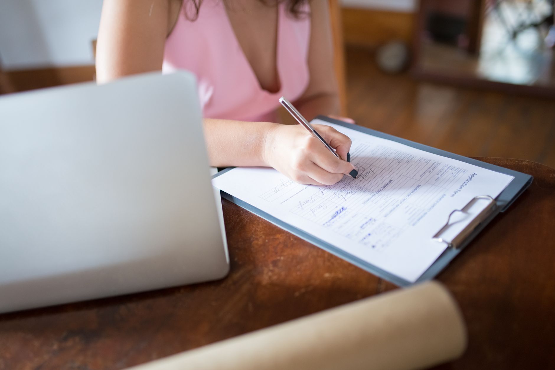 A woman writes on a document clipped to a clipboard, with a laptop nearby on a wooden desk.