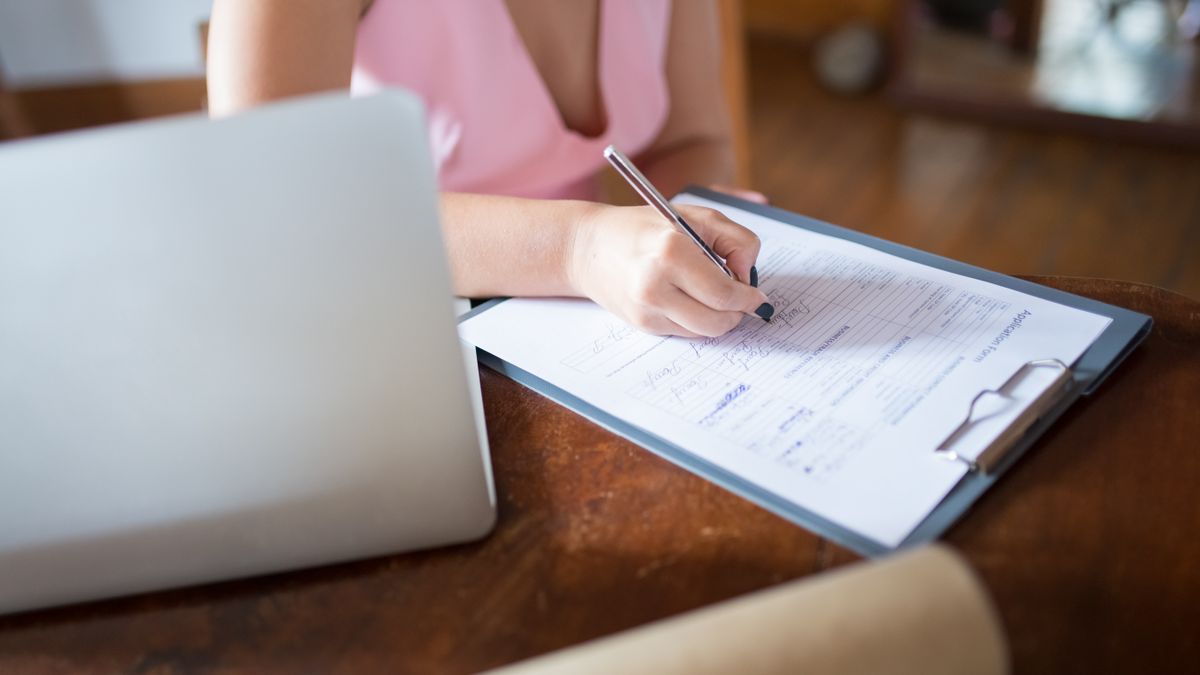 A woman writes on a document clipped to a clipboard, with a laptop nearby on a wooden desk.
