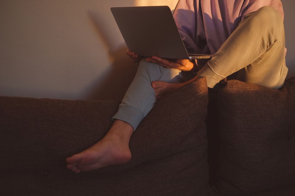 A person sitting barefoot on a couch, using a laptop in warm lighting, indoors.