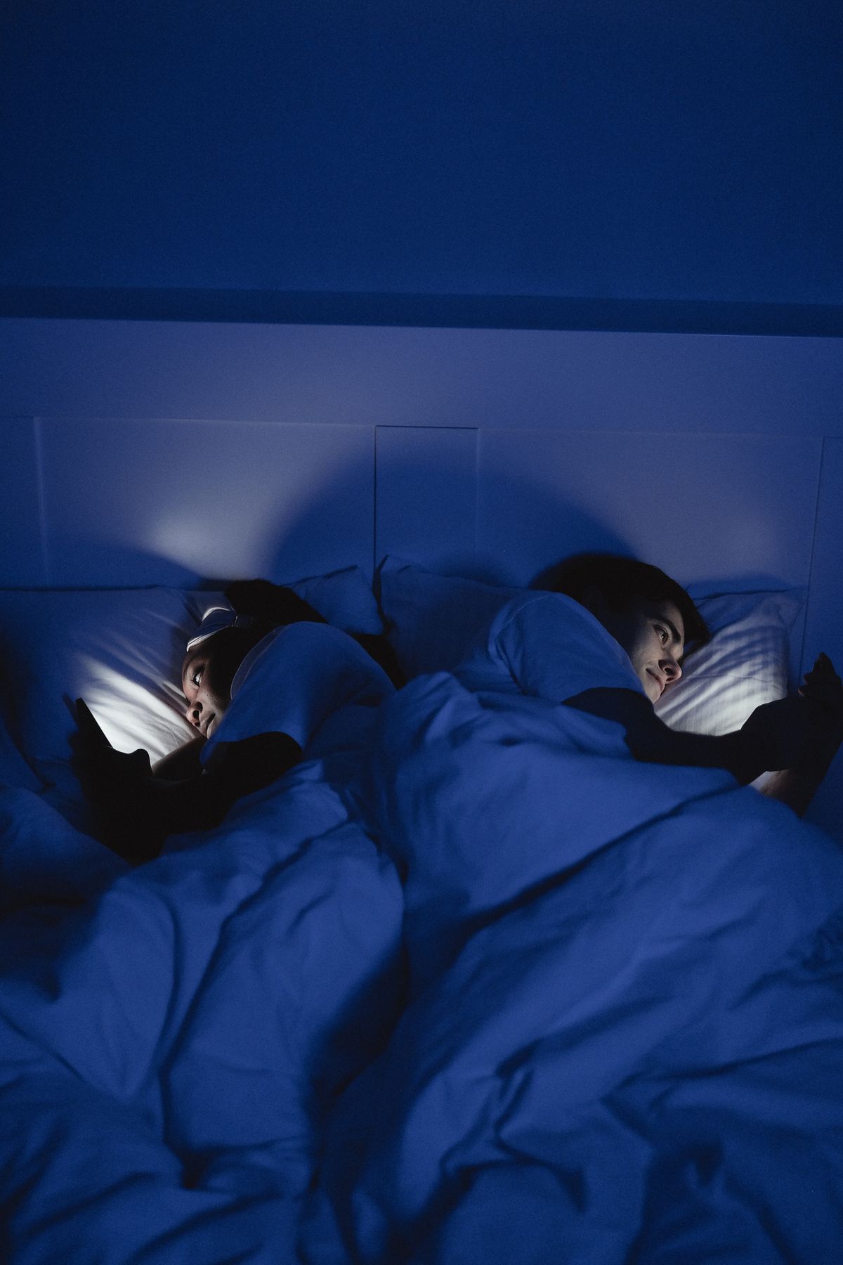 Two people in bed using smartphones under blue-toned light at night