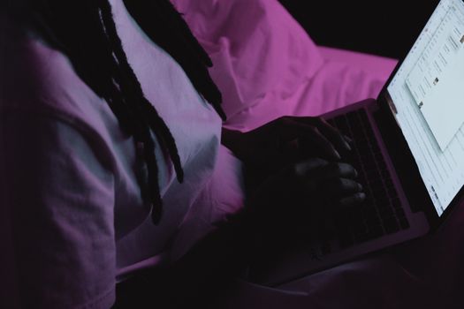 A woman with dreadlocks typing on a laptop in a dimly lit room at night.