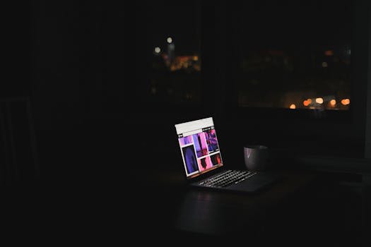 A laptop glows in a dark room at night with a cityscape through the window. Ideal for tech and solitude themes.