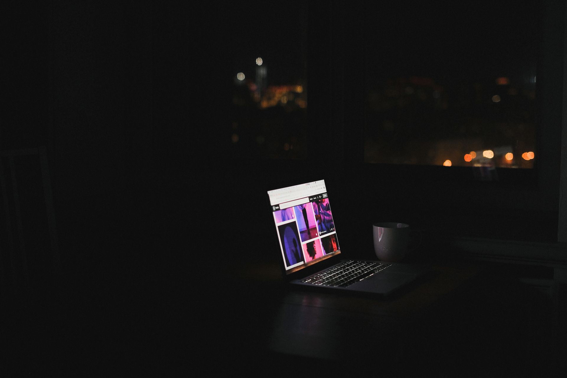 A laptop glowing in a dark room at night with a city skyline through the window.
