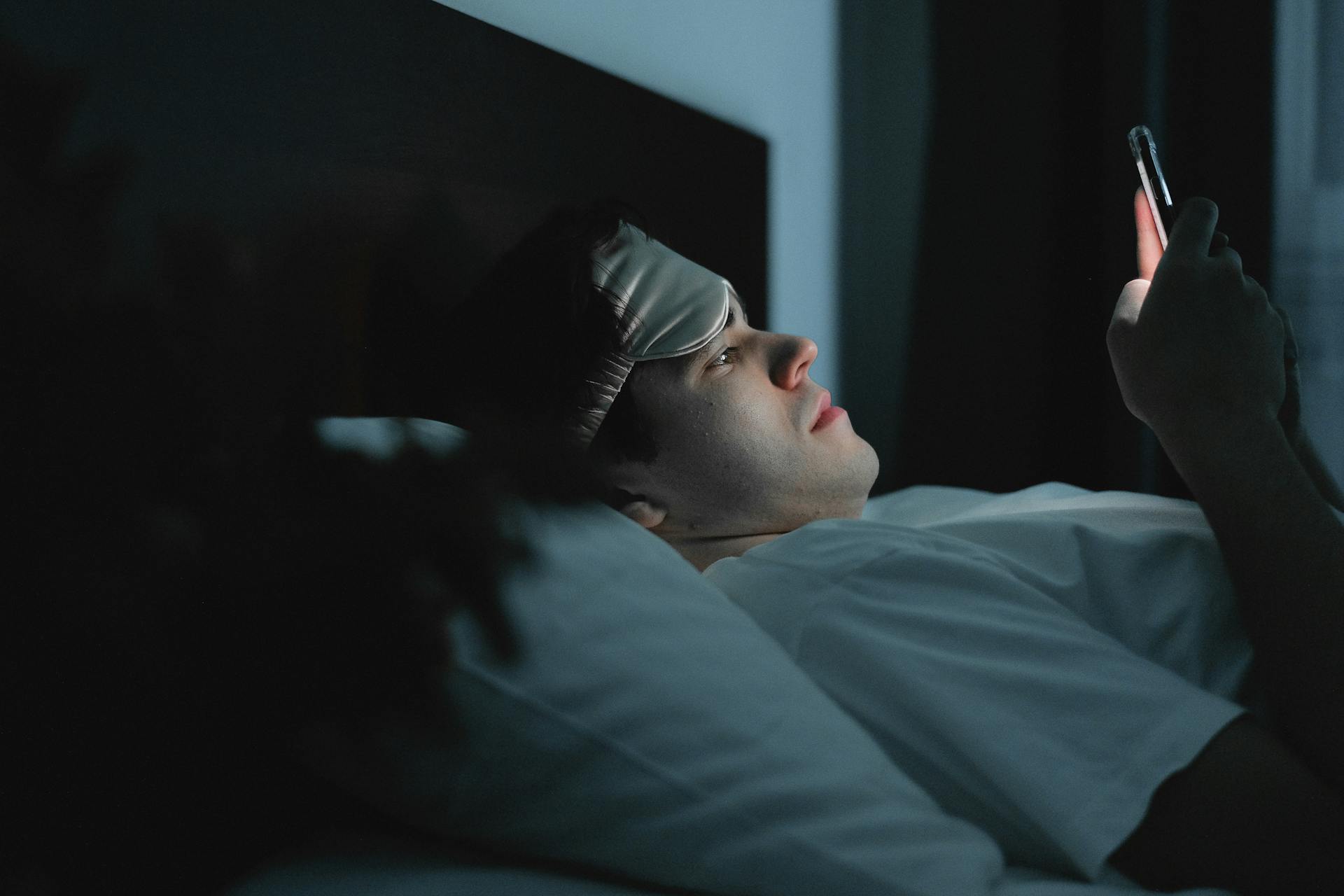 A man wearing a sleep mask late at night using a smartphone on his bed.