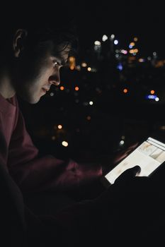 Side view of a man using a digital tablet in the dark outdoors, with city lights in the background.