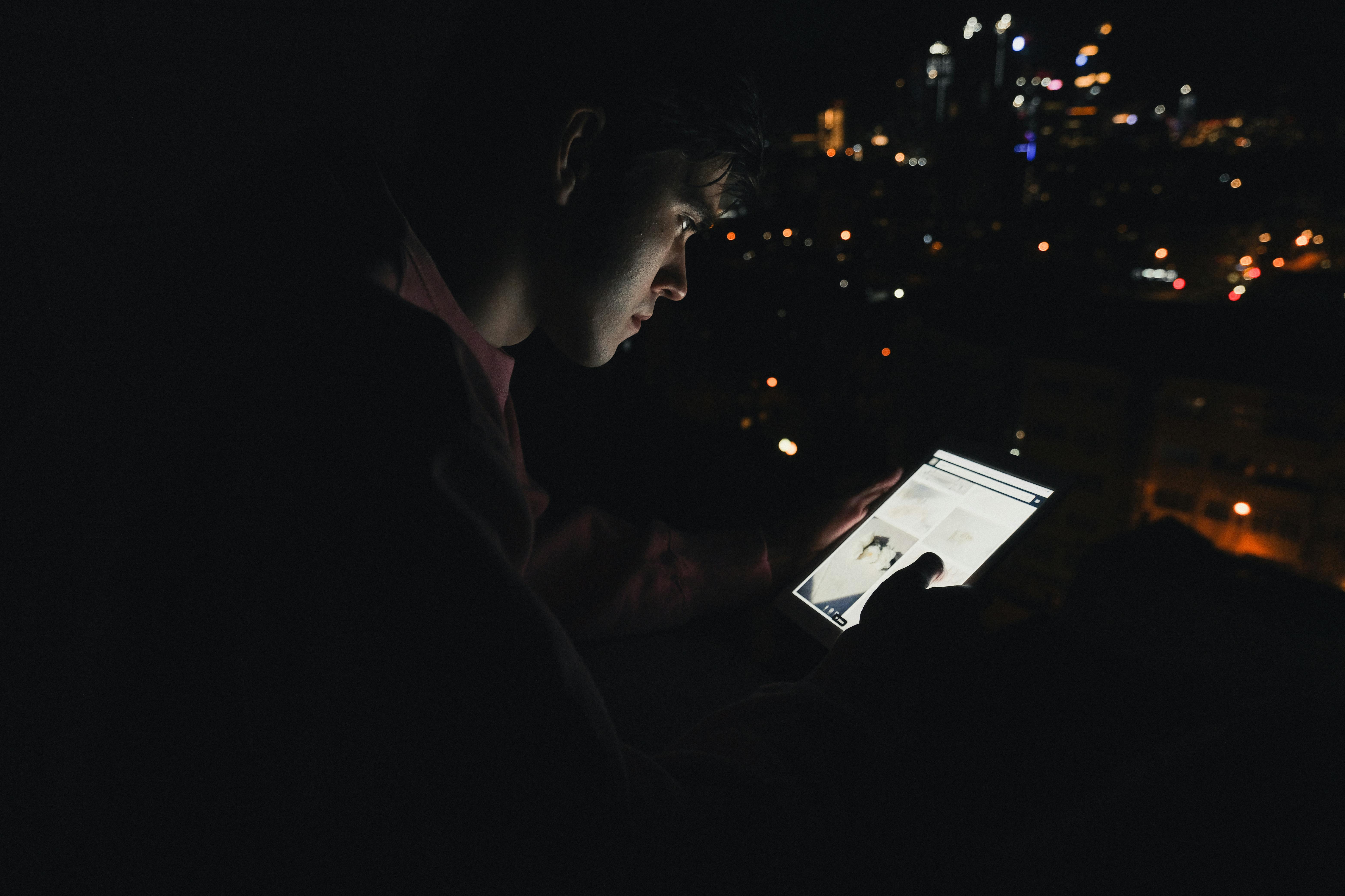 Silhouette of a man using a glowing digital tablet with city lights in the background at night.