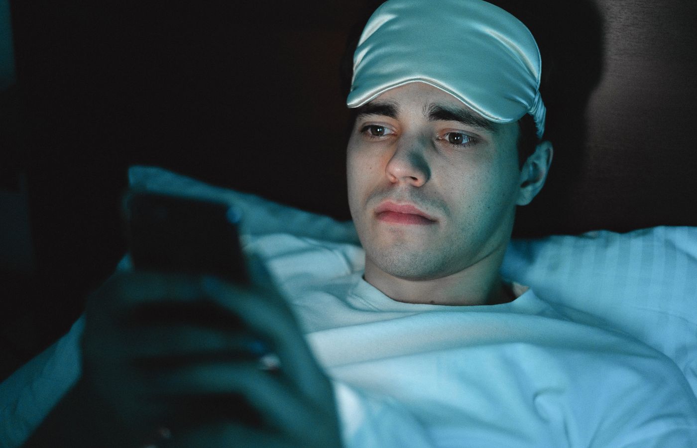 A man wearing a sleeping mask uses his smartphone while lying in bed at night.