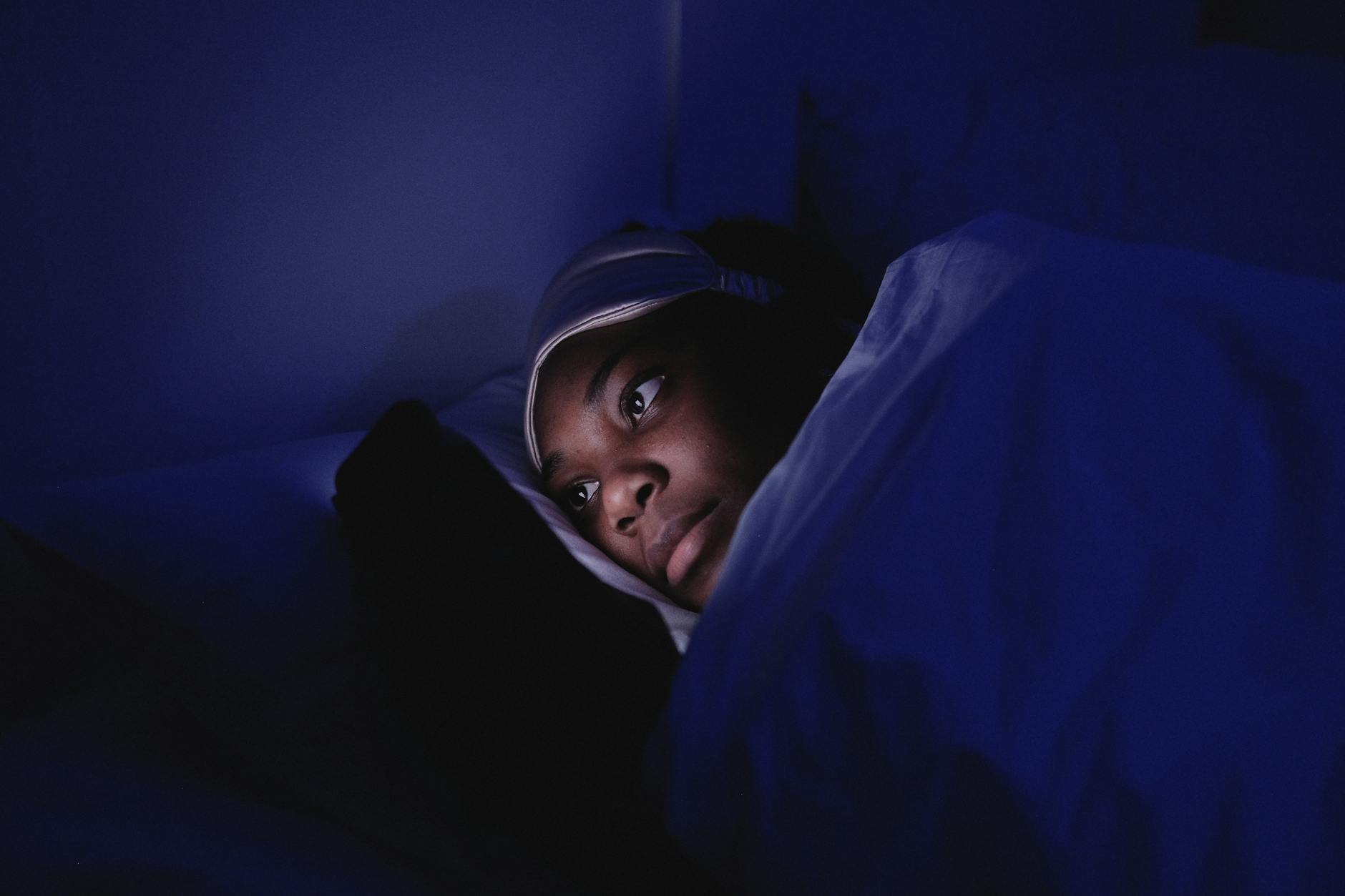 A woman lies in bed at night using her smartphone, illuminated by its screen.