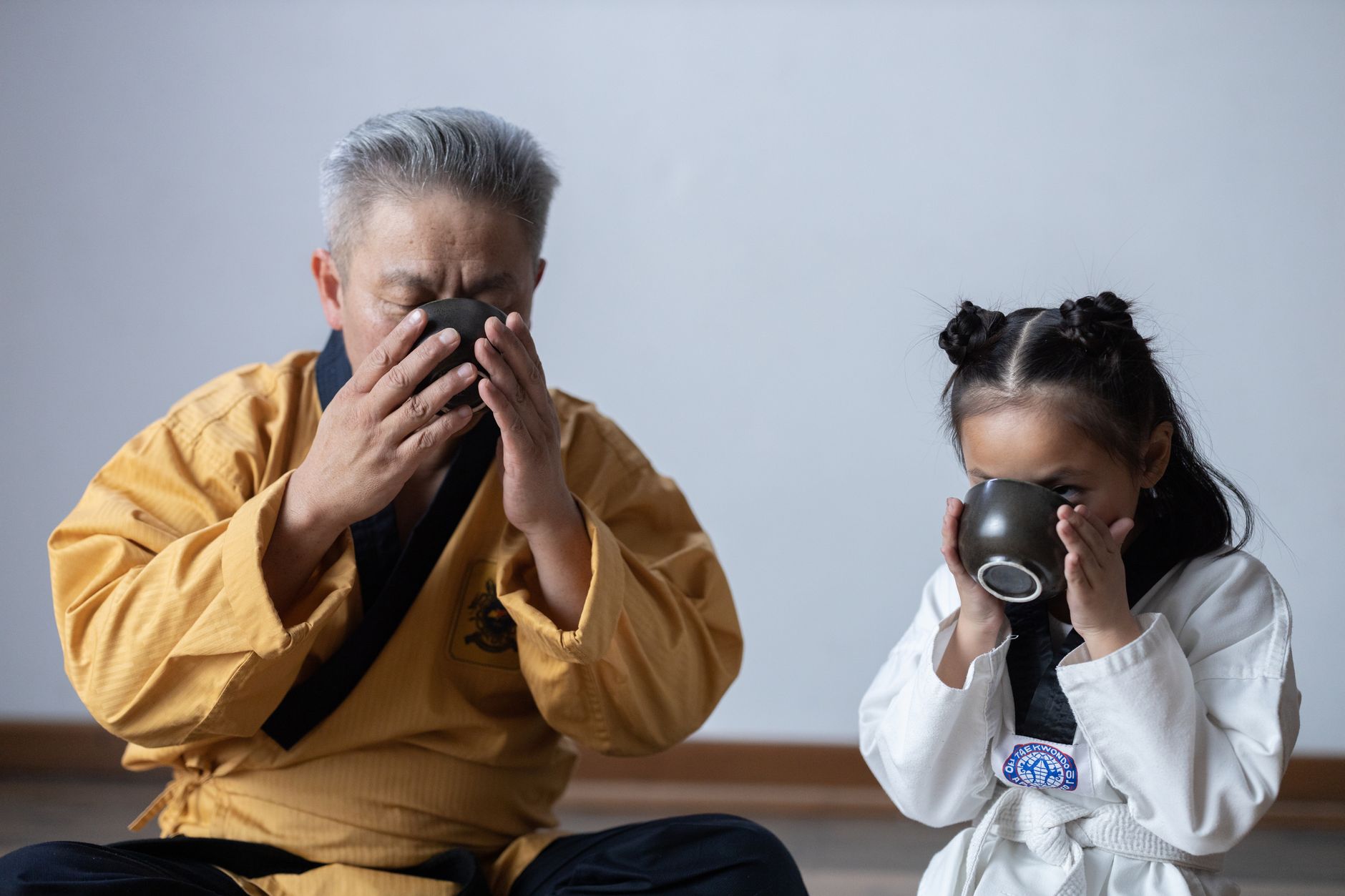 Elderly man and young girl in martial arts attire enjoy a traditional tea ceremony indoors.