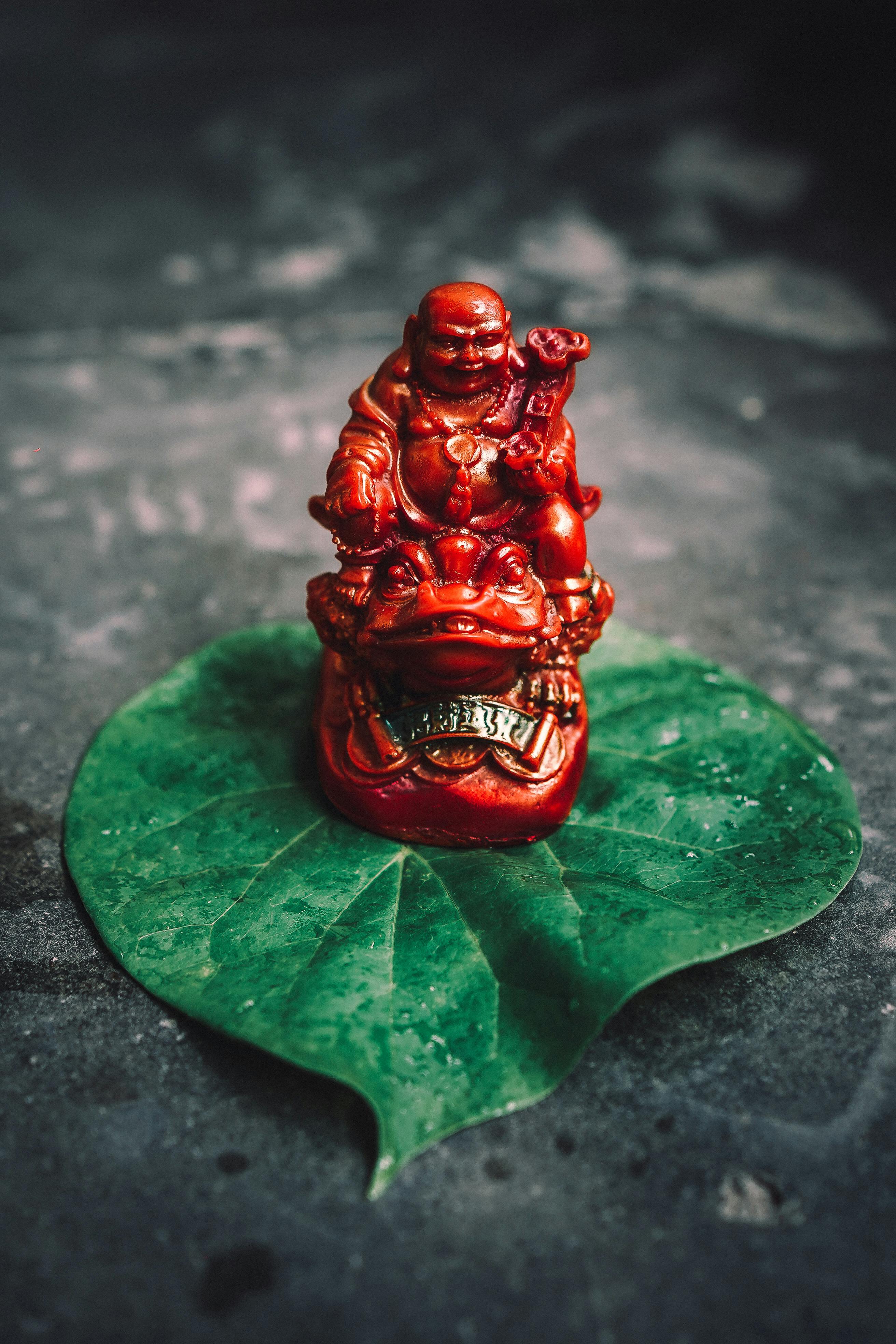 A red Buddha figurine on a green leaf with water droplets, captured in a close-up shot.