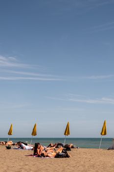Anonymous tourists resting on sandy shore against row of sunshades and ocean under cloudy blue sky