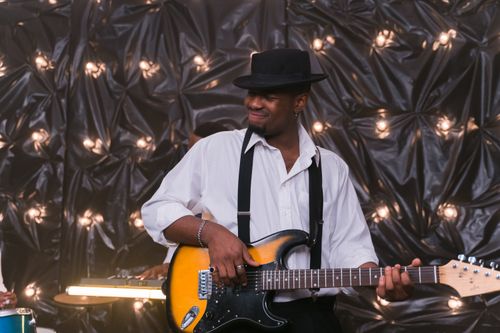 Free African American guitarist wearing a fedora, performing with electric guitar in festive setting. Stock Photo