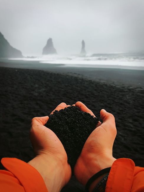 Close-up of hands holding black sand with Reynisfjara Beach's iconic sea stacks in the background.