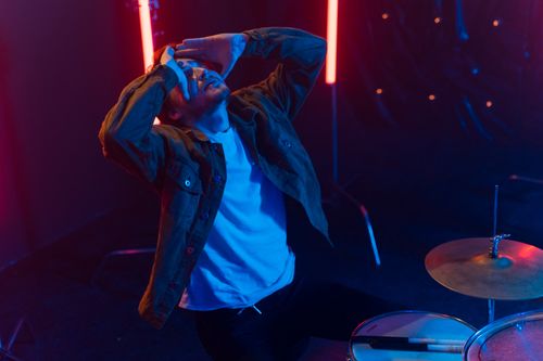 Free A man in a denim jacket showing frustration while seated at a drum set under neon lights. Stock Photo