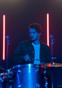 A male musician in a denim jacket playing drums under vibrant neon lights.