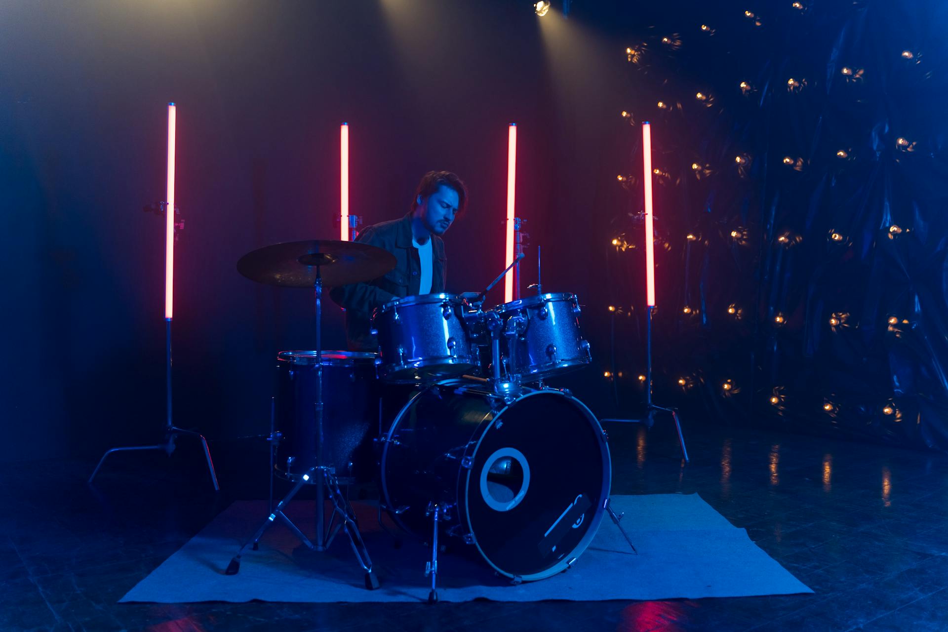 A musician plays drums under dramatic neon lights in a dark studio setting.