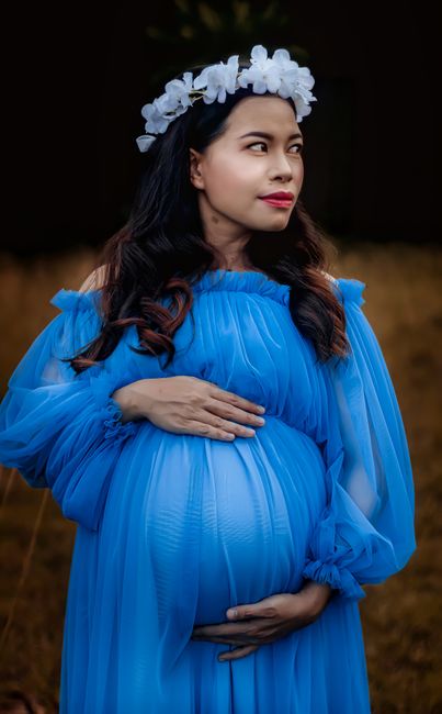 Thoughtful pregnant Asian female with dark hair in elegant blue dress and floral wreath touching belly and looking away in nature in daytime