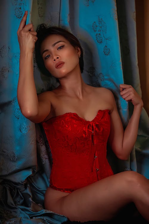 Charming Asian woman in red corset sitting near blue curtain
