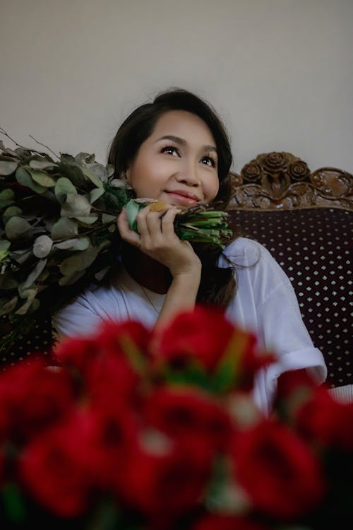 Dreamy Asian female with dark hair in casual clothes sitting with bouquet of flowers in hand in light room and looking away