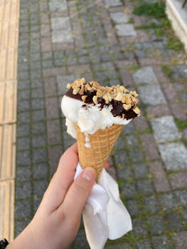 Hand holding a melting ice cream cone with chocolate and nut topping outdoors.