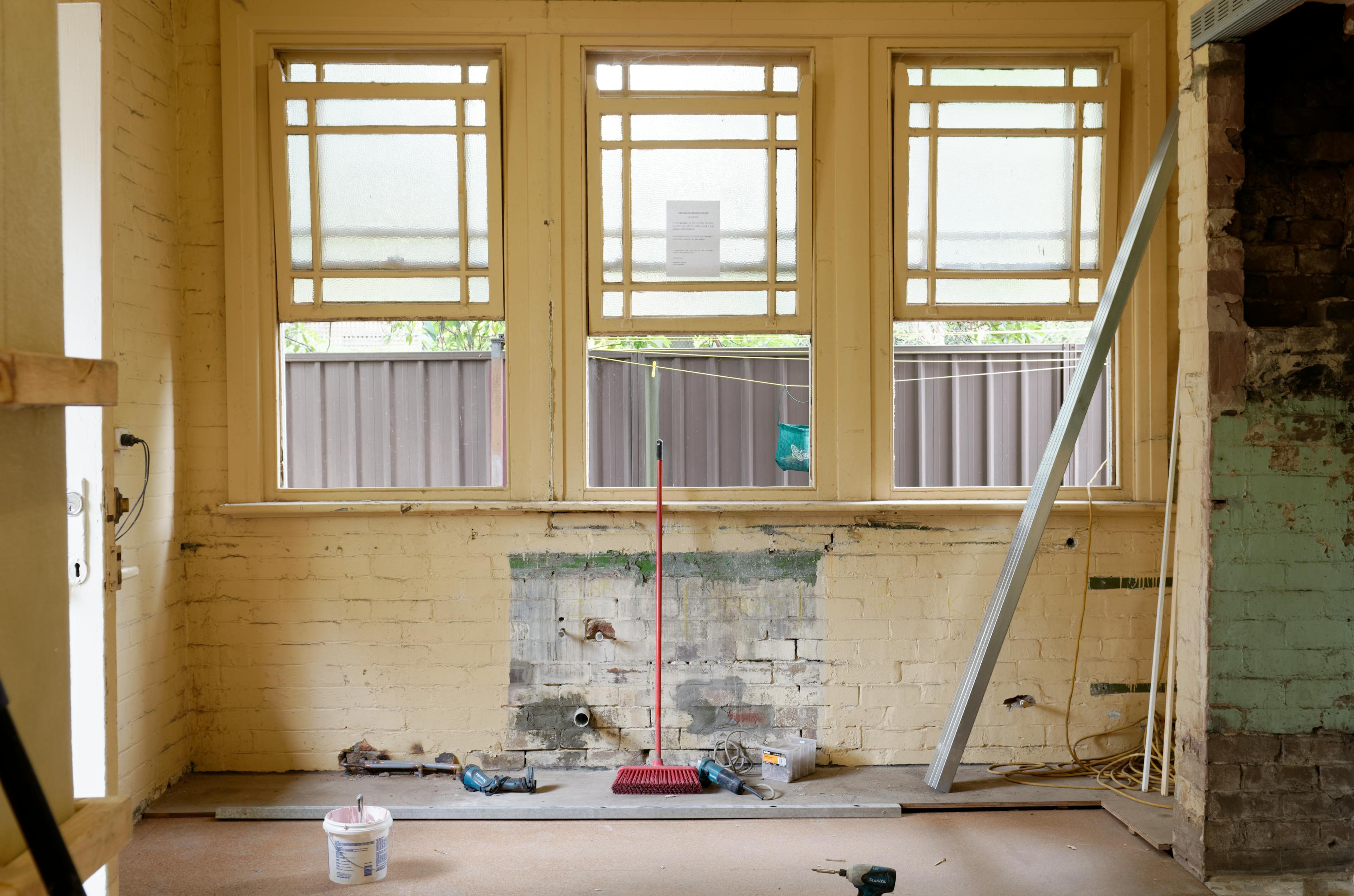Interior wall of an apartment undergoing renovations that has been stripped down to the brick; a broom and other construction tools sit beneath open windows.