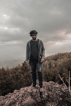 一名男子穿着休闲装，站在岩石山上，背景是多云的美景