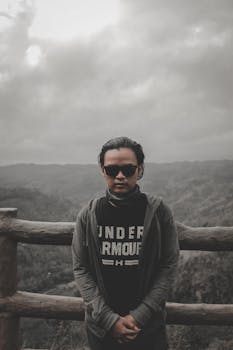 A man stands confidently wearing sunglasses against a cloudy mountain backdrop.