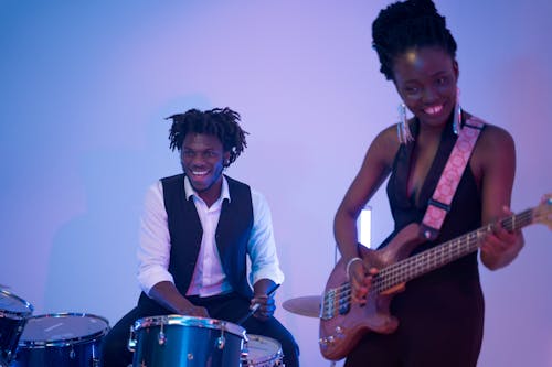 Man in White Button Up Shirt Playing Drums Beside a Woman Playing Electric Guitar